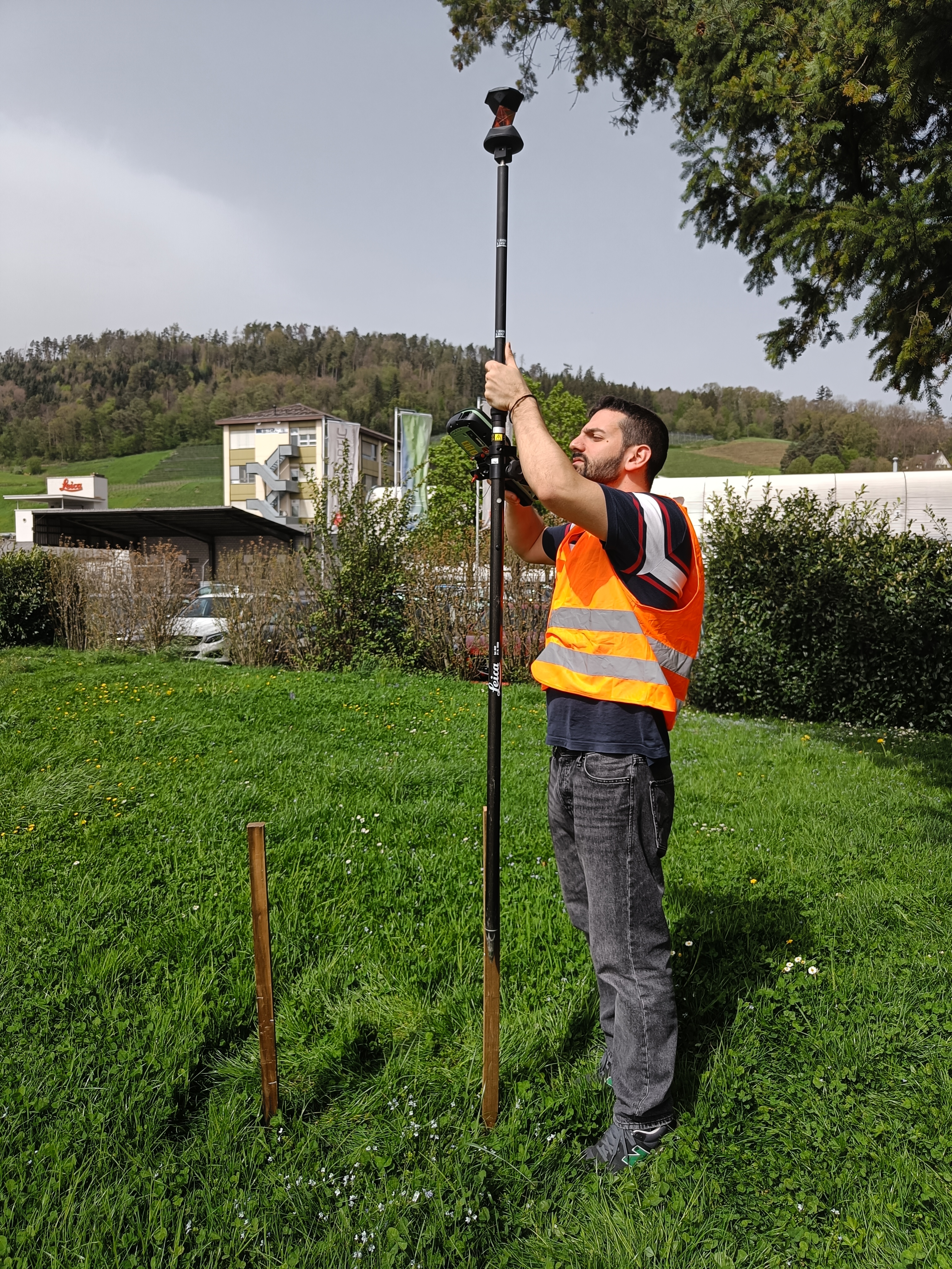 A surveyor in an orange safety vest hold AP20 AutoPole vertically in a grassy area with hills and buildings in the background.